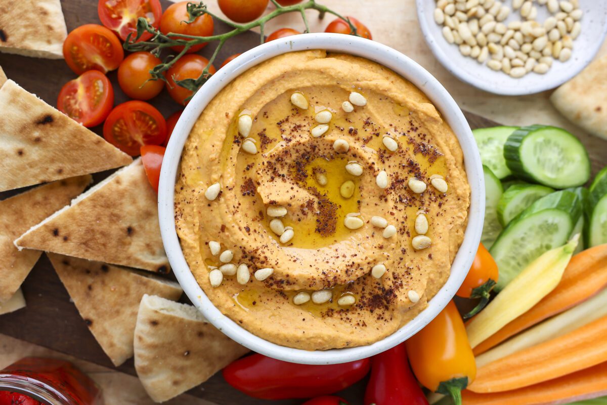 hummus in small white serving bowl topped with sumac, olive oil and pine nuts. vegetables and pita triangles arranged around serving bowl