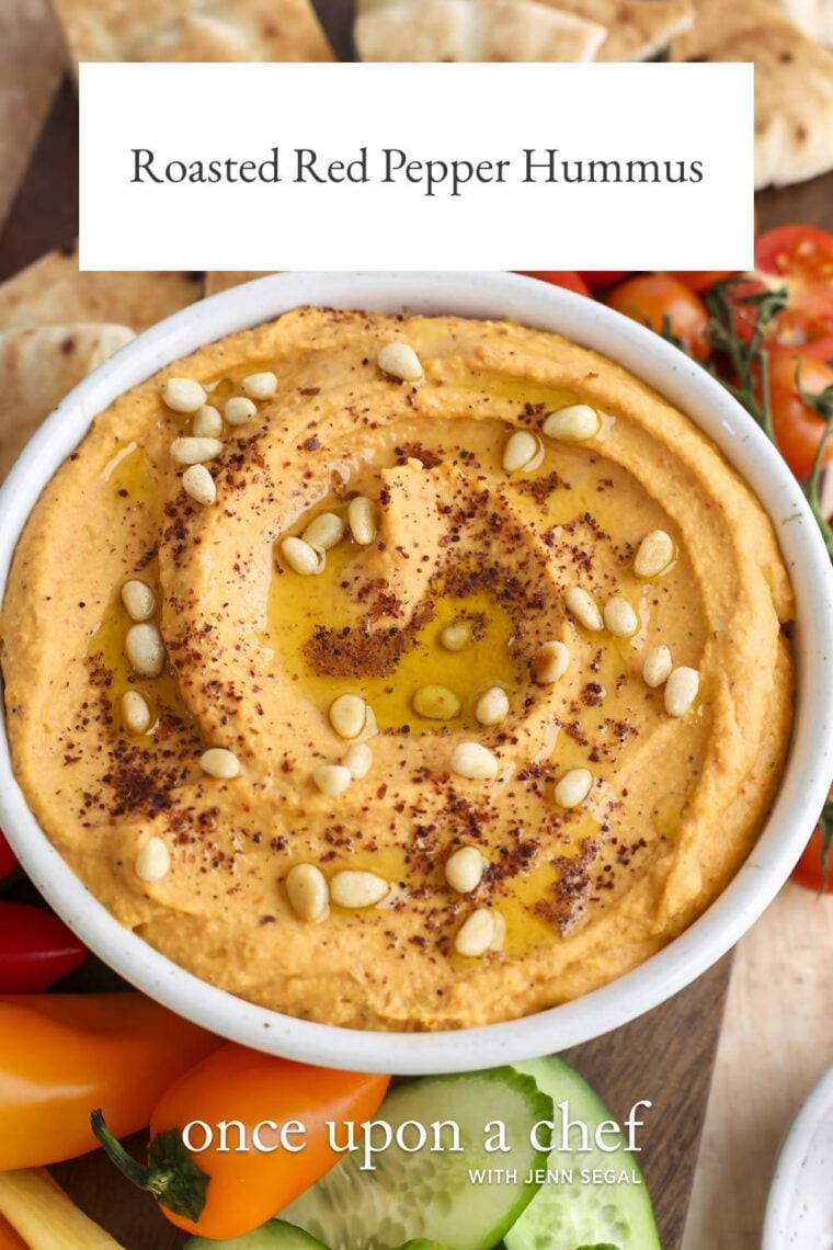 hummus topped with pine nuts, sumac, and olive oil in white serving bowl; bowl is placed between fresh vegetables and pita triangles