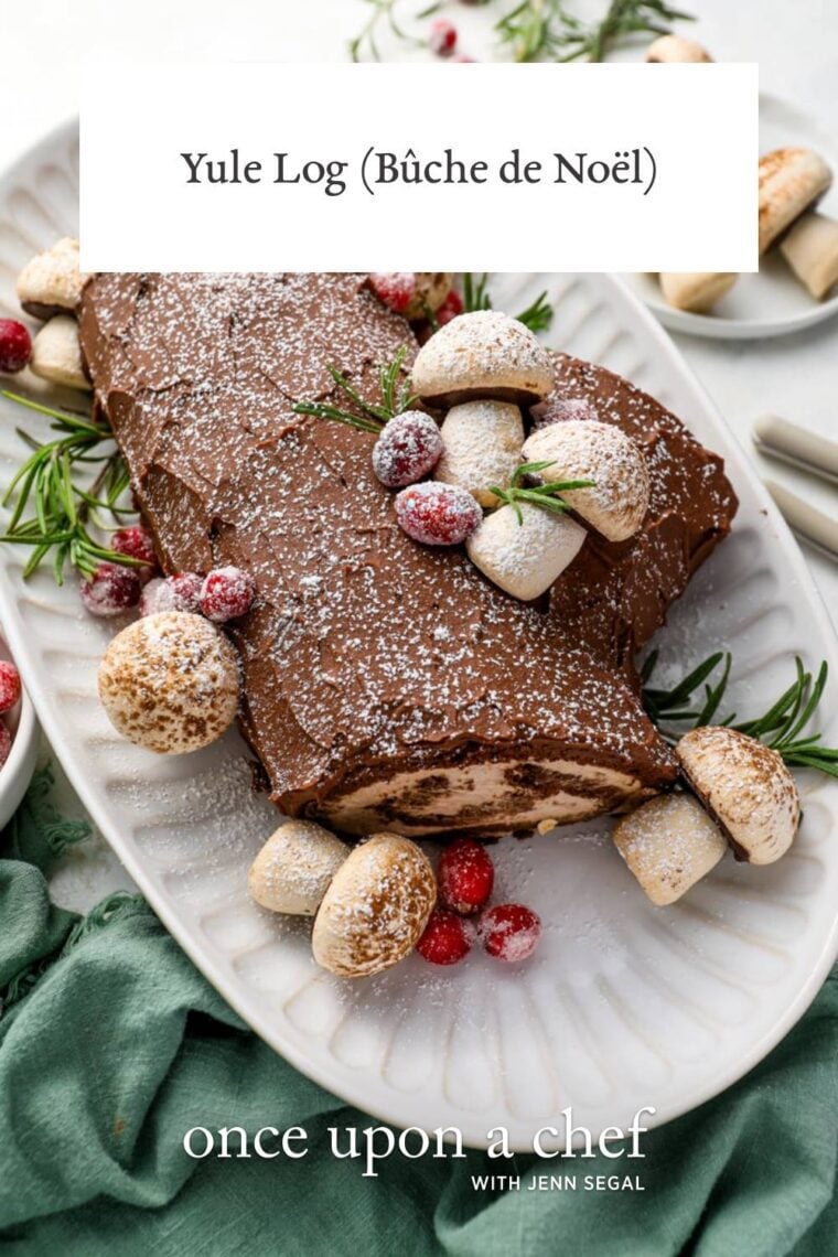 white serving platter holding yule log cake adorned with meringue mushrooms, sugared cranberries, and sprigs of rosemary