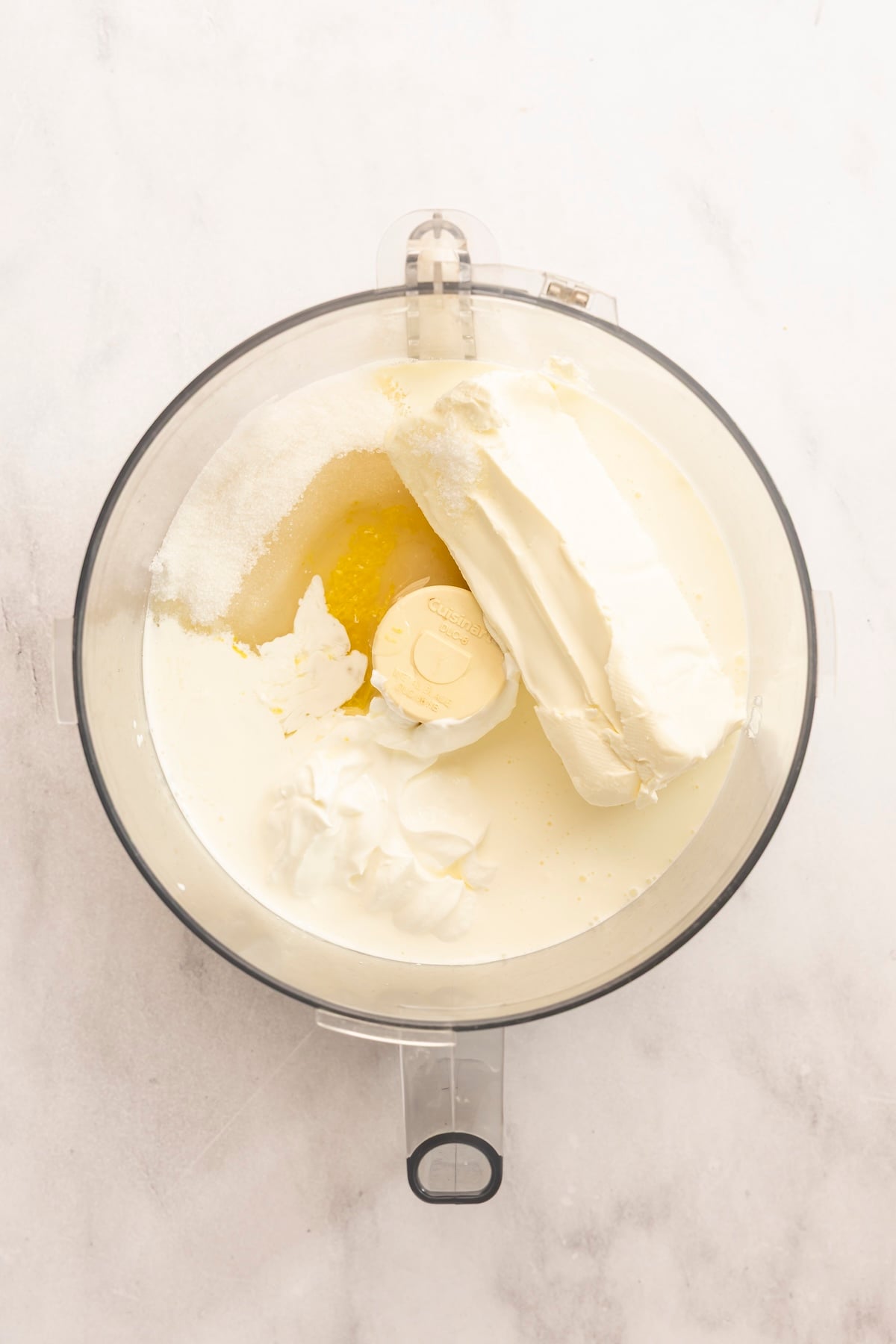 food processor with heavy cream, cream cheese, sour cream, granulated sugar, lemon zest, lemon juice, and salt added to it