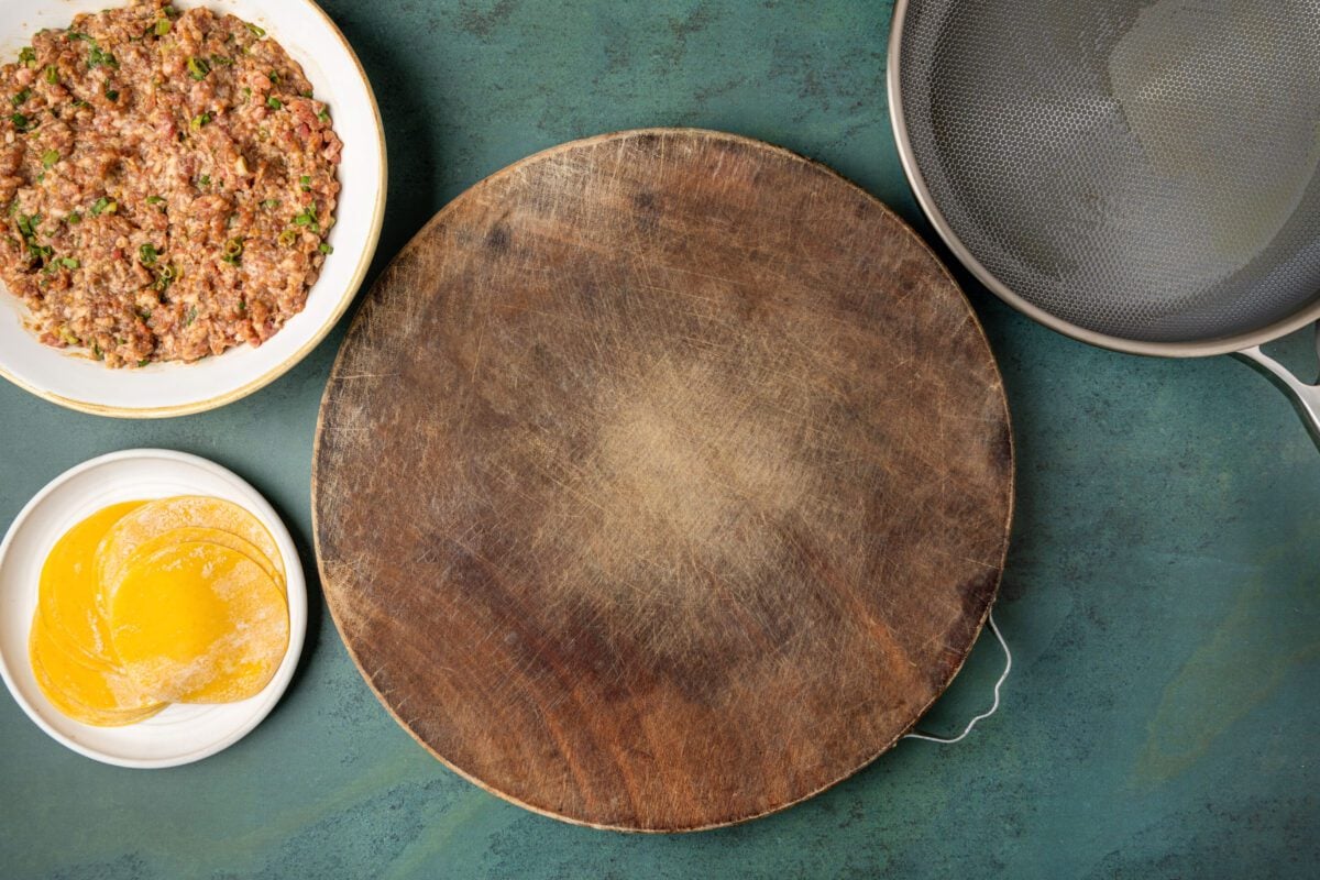 assembly station with ground pork mixture, dumpling wrappers, wooden work surface, and non-stick pan