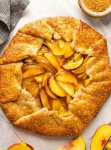 Peach galette on parchment with sliced peaches and bowl of turbinado sugar nearby.