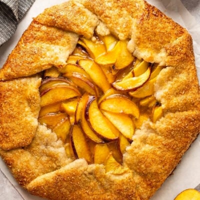 Peach galette on parchment with sliced peaches and bowl of turbinado sugar nearby.