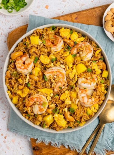Bowl of pineapple fried rice with small bowls of scallions and cashews.